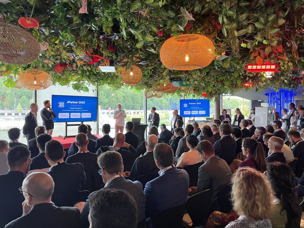 Photo d’un événement professionnel avec une conférence devant un public, écrans affichant “JPartner 2025”, dans un espace décoré de végétation suspendue.
